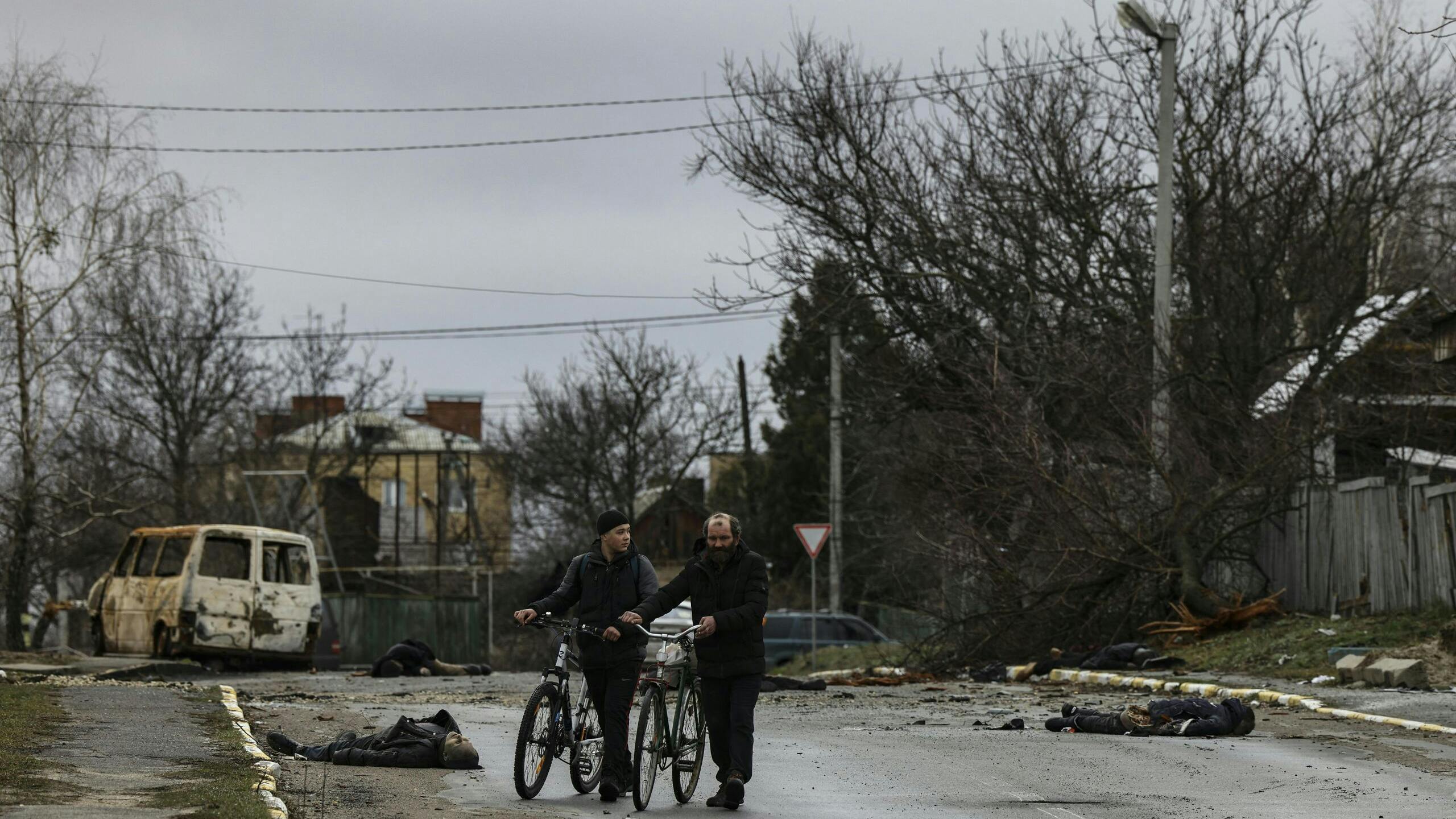 Twee personen duwen hun fiets terwijl ze een straat oversteken waar verschillende dode lichamen op de grond liggen in Boetsja.