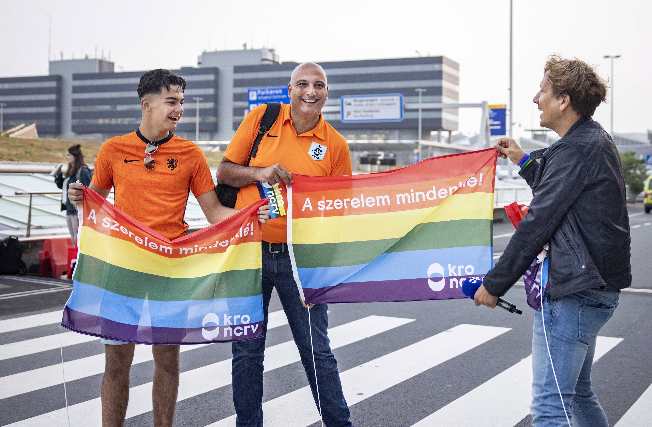 KRO-NCRV-presentator Klaas van Kruistum deelde op Schiphol regenboogvlaggen uit aan voetbalsupporters die naar de wedstrijd in Hongarije gingen.