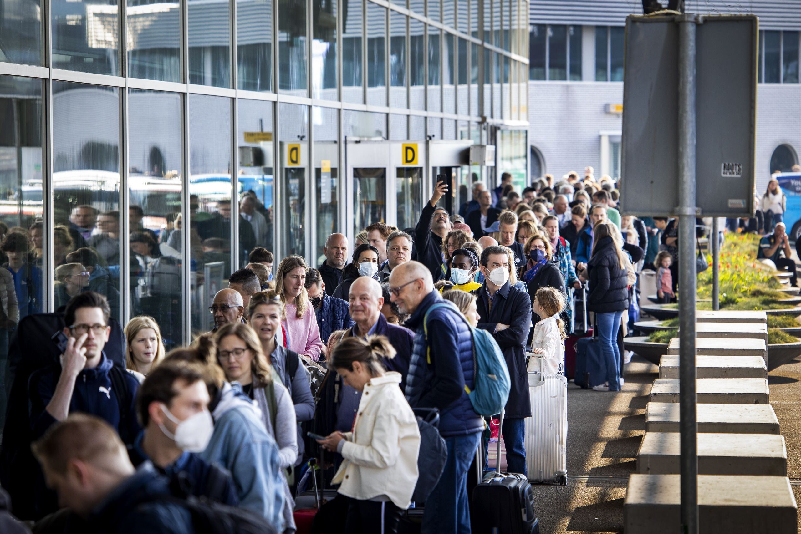 Reizigers staan buiten in de rij bij een vertrekhal van Schiphol.