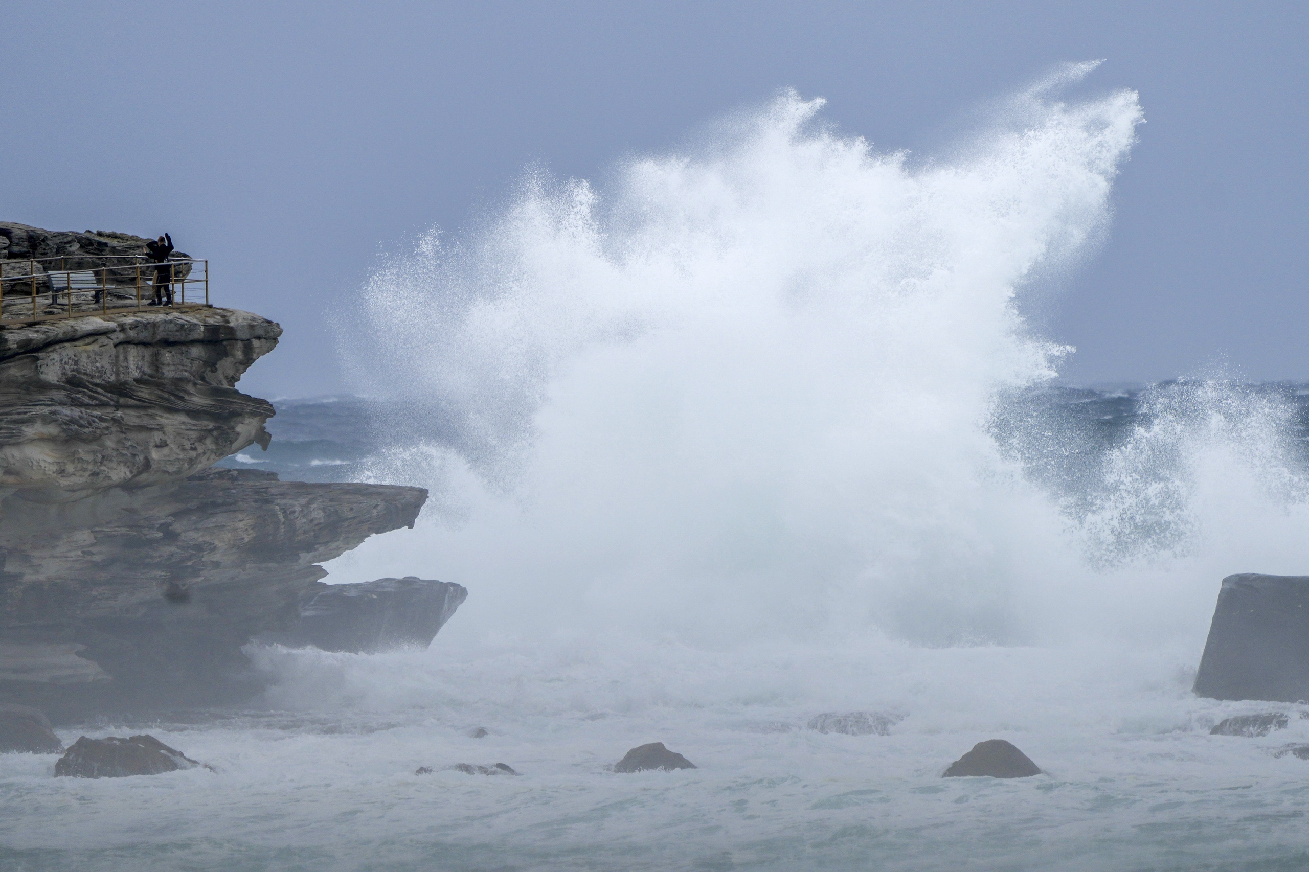 People stand on a rock ledge as huge swells hit the headland at Bondi Beach in Sydney, Australia, Sunday, July 3, 2022. A severe weather warning for heavy rainfall and strong winds has been issued for Sydney, as parts of NSW have received more than their monthly average rainfall within hours this weekend. (AP Photo/Mark Baker)